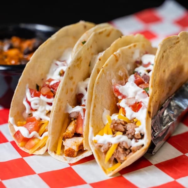 Three Fat Floyd's Barbecue tacos with meat, cheese, tomatoes, and sour cream on a checkered paper.